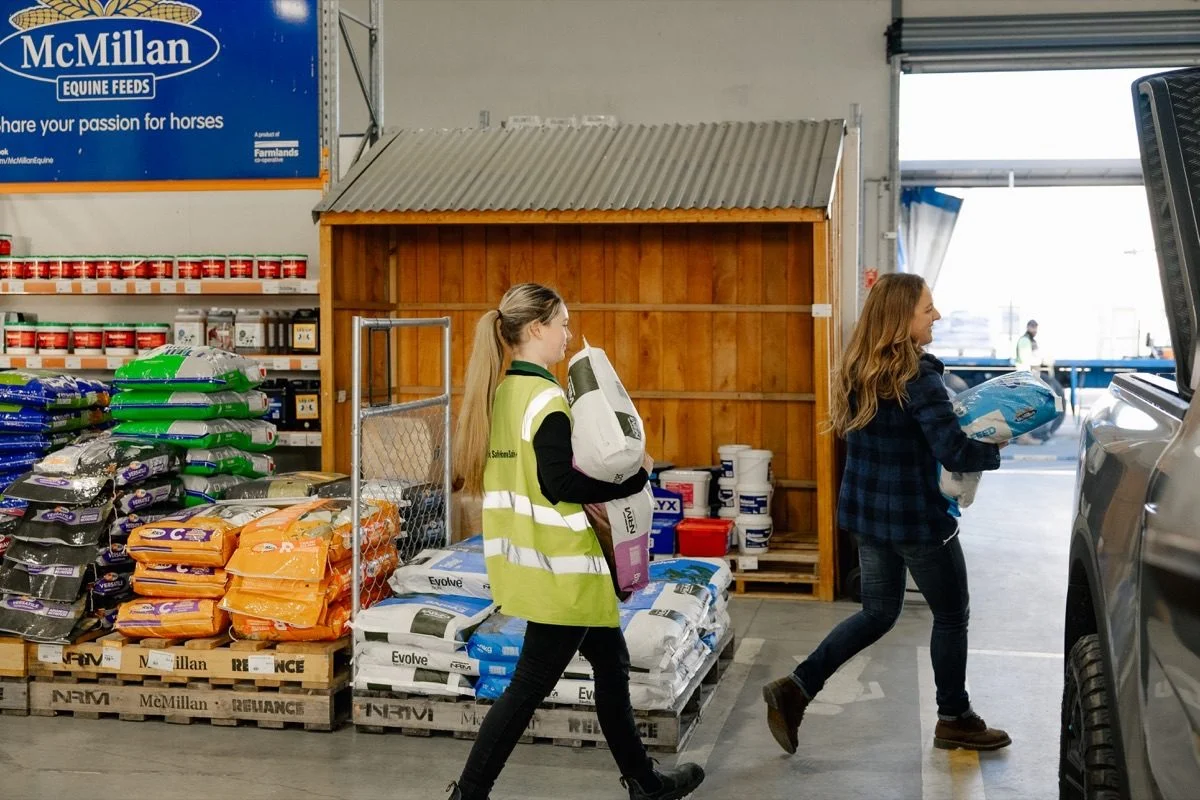 Shareholder benefits: Two people carrying animal feed bags in a Farmlands store with shelves of more feed.