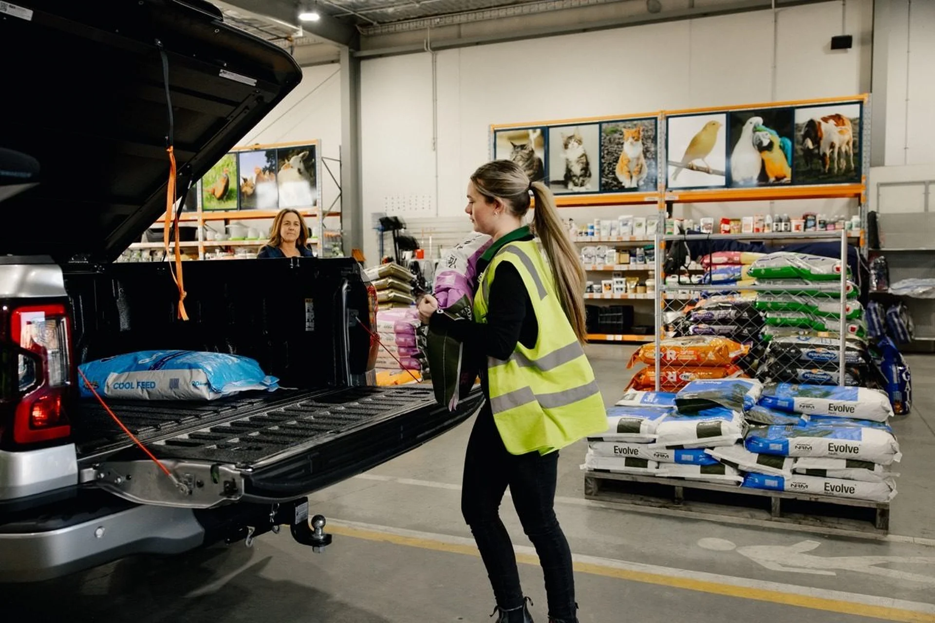 Shop with Farmlands: A person in a high-visibility vest loads pet food bags into a truck at a store.