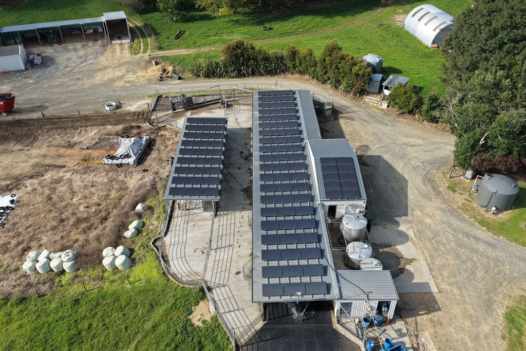 Aerial view of farm buildings with solar panels on the roofs, surrounded by green fields, trees, and farm equipment.