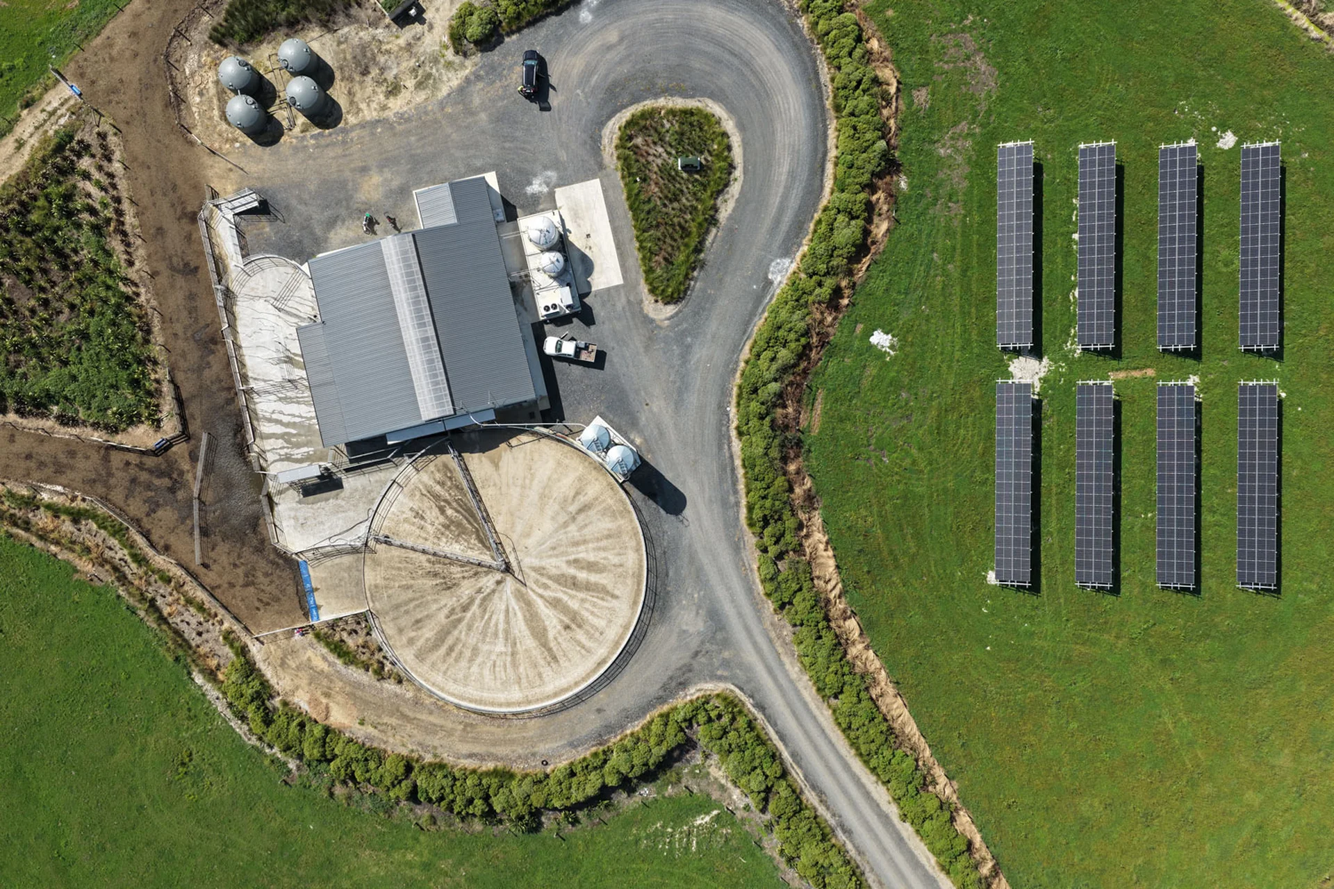 Aerial view of a farm facility with a large round silo, several tanks, trucks, and an array of solar panels on a green field.