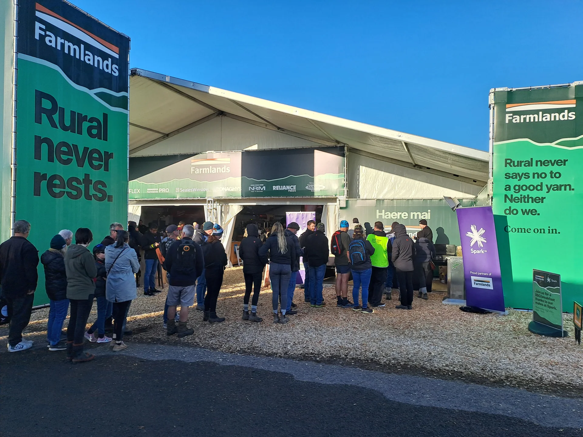 People queue outside a large tent at an outdoor event. Banners with "Farmlands" and rural-themed slogans flank the entrance.