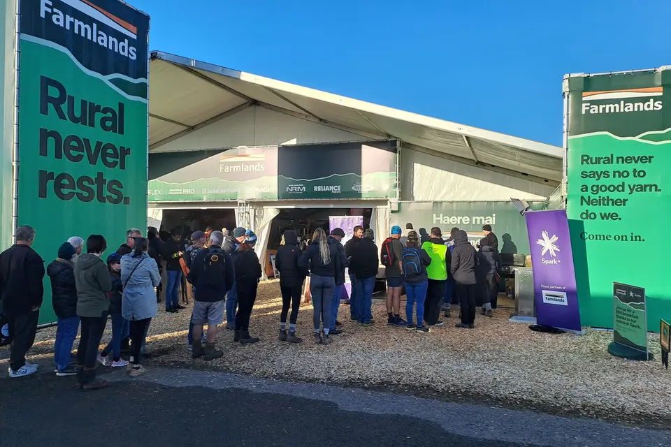 People queue outside a large tent at an outdoor event. Banners with "Farmlands" and rural-themed slogans flank the entrance.