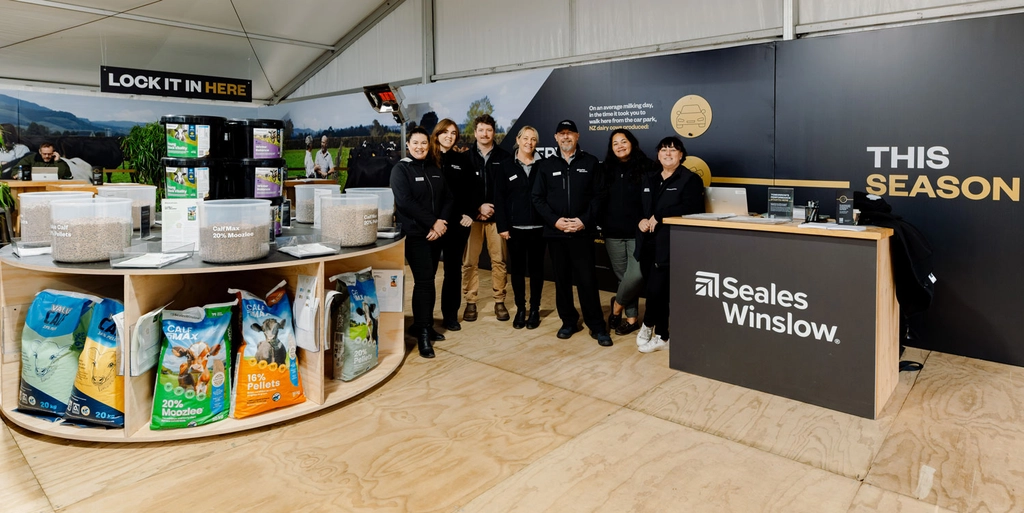 A group of eight people in matching dark jackets stands at a Fieldays booth featuring SealesWinslow animal feed products.