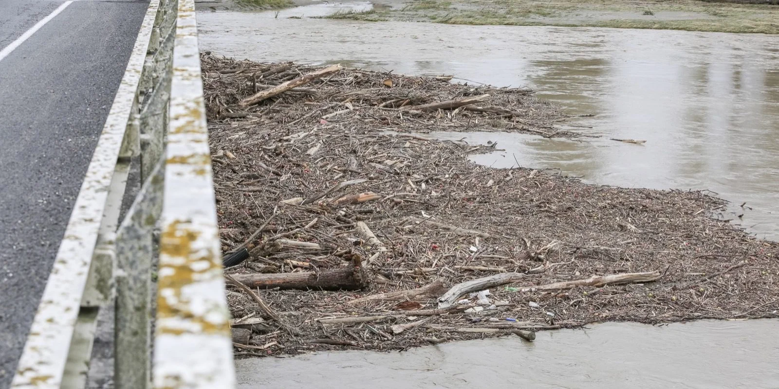 Rural insurance claim advice: Flooded river with debris accumulation by a road bridge, showing muddy water and scattered branches.