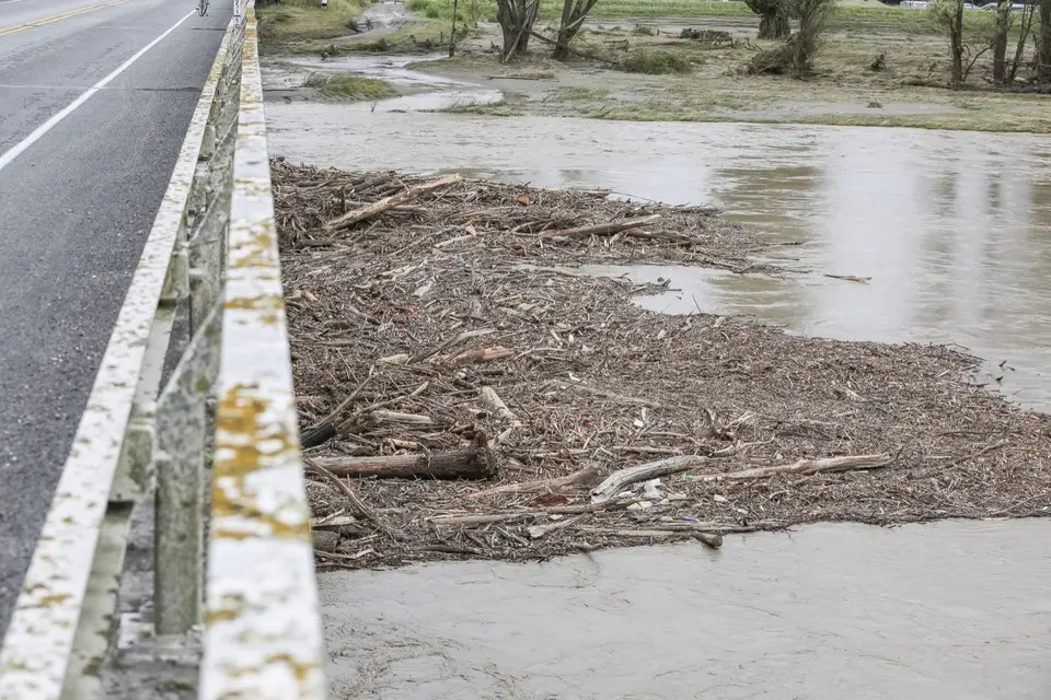 Rural insurance claim advice: Flooded river with debris accumulation by a road bridge, showing muddy water and scattered branches.