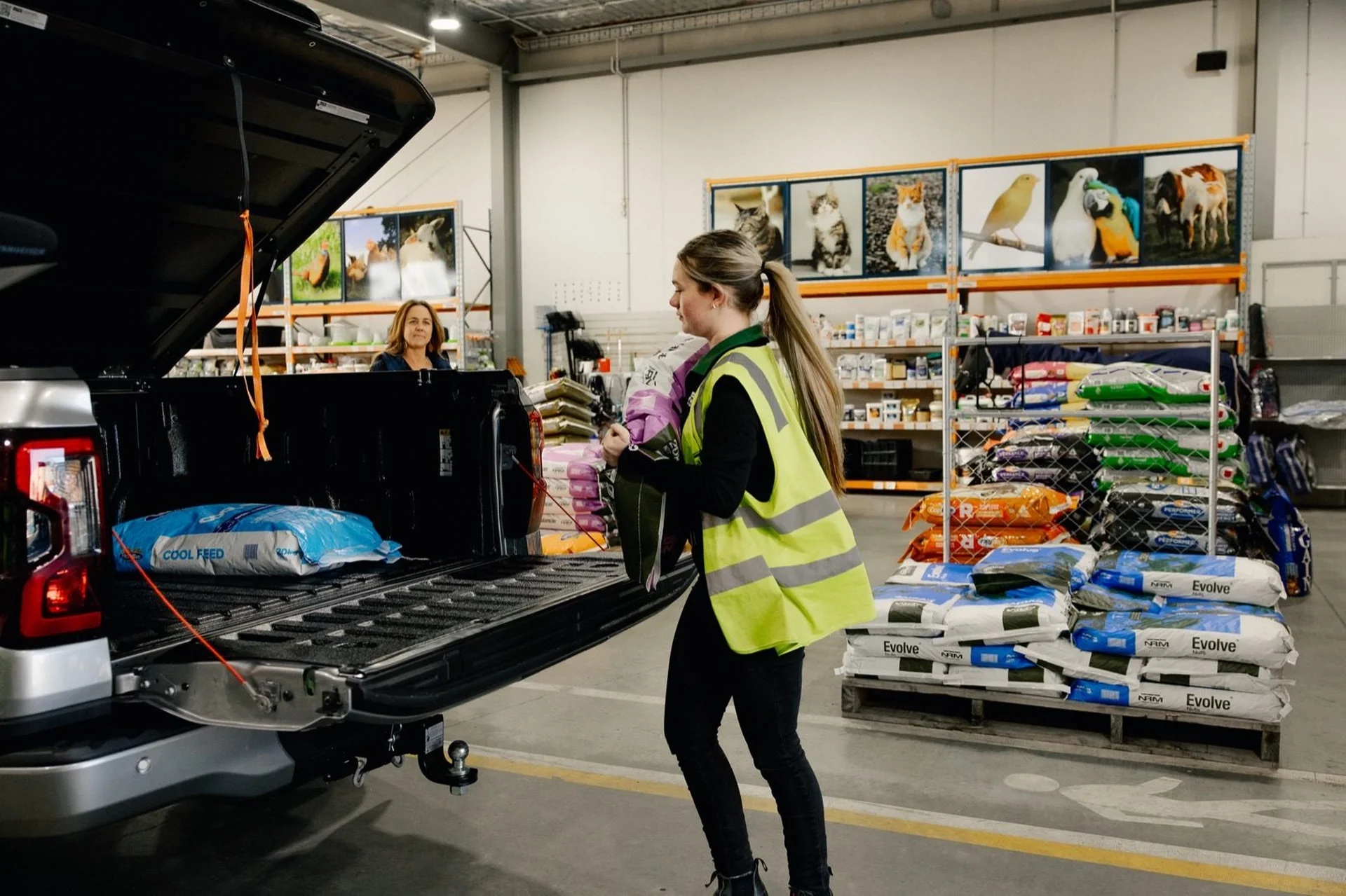 Farm Flexi: Worker in a safety vest loads pet supplies into a truck at a store with shelves of animal products in the background.