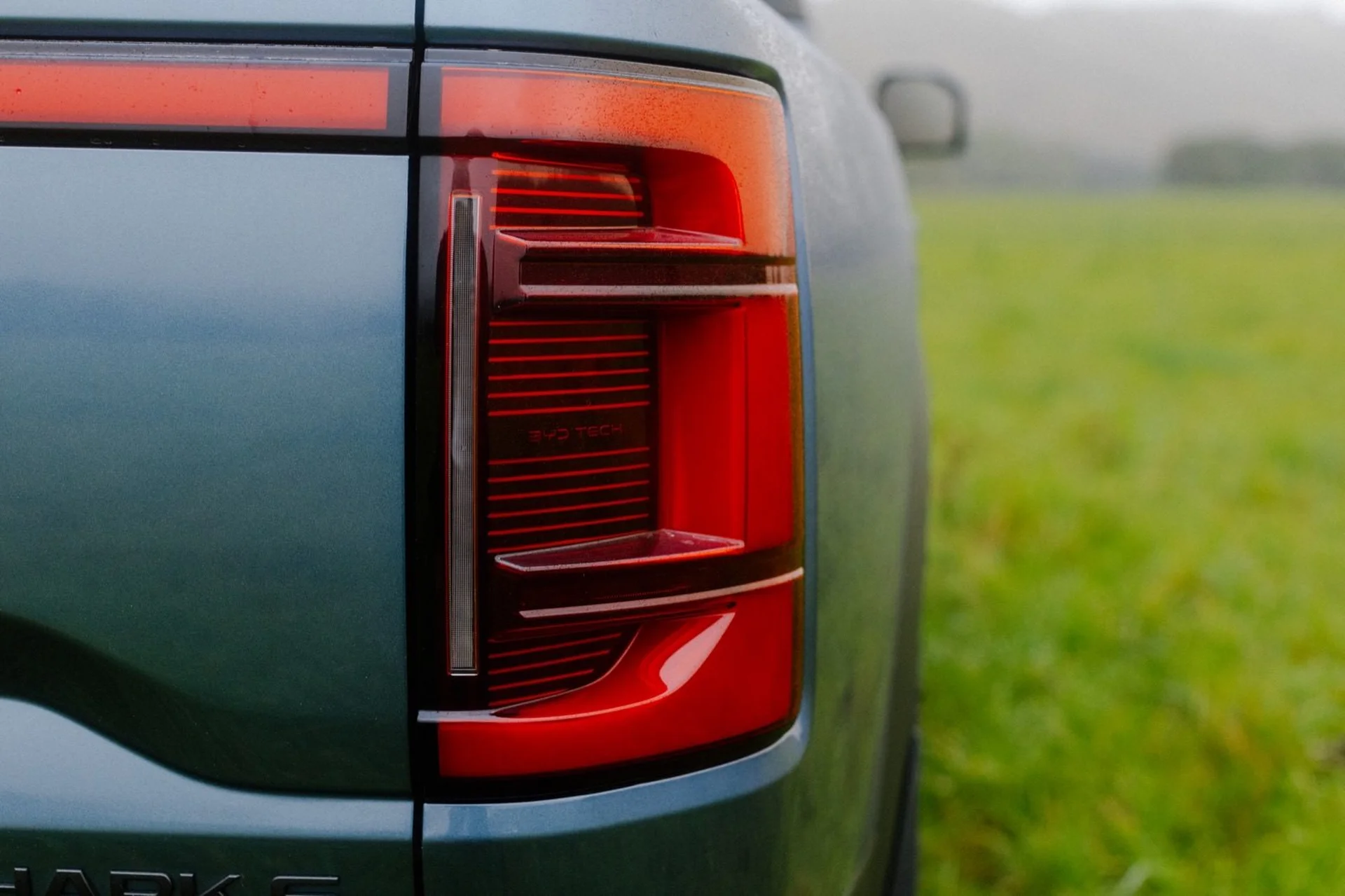 Farm Fixed: Close-up of a blue vehicle's rear left taillight with red details and a blurred green field in the background.