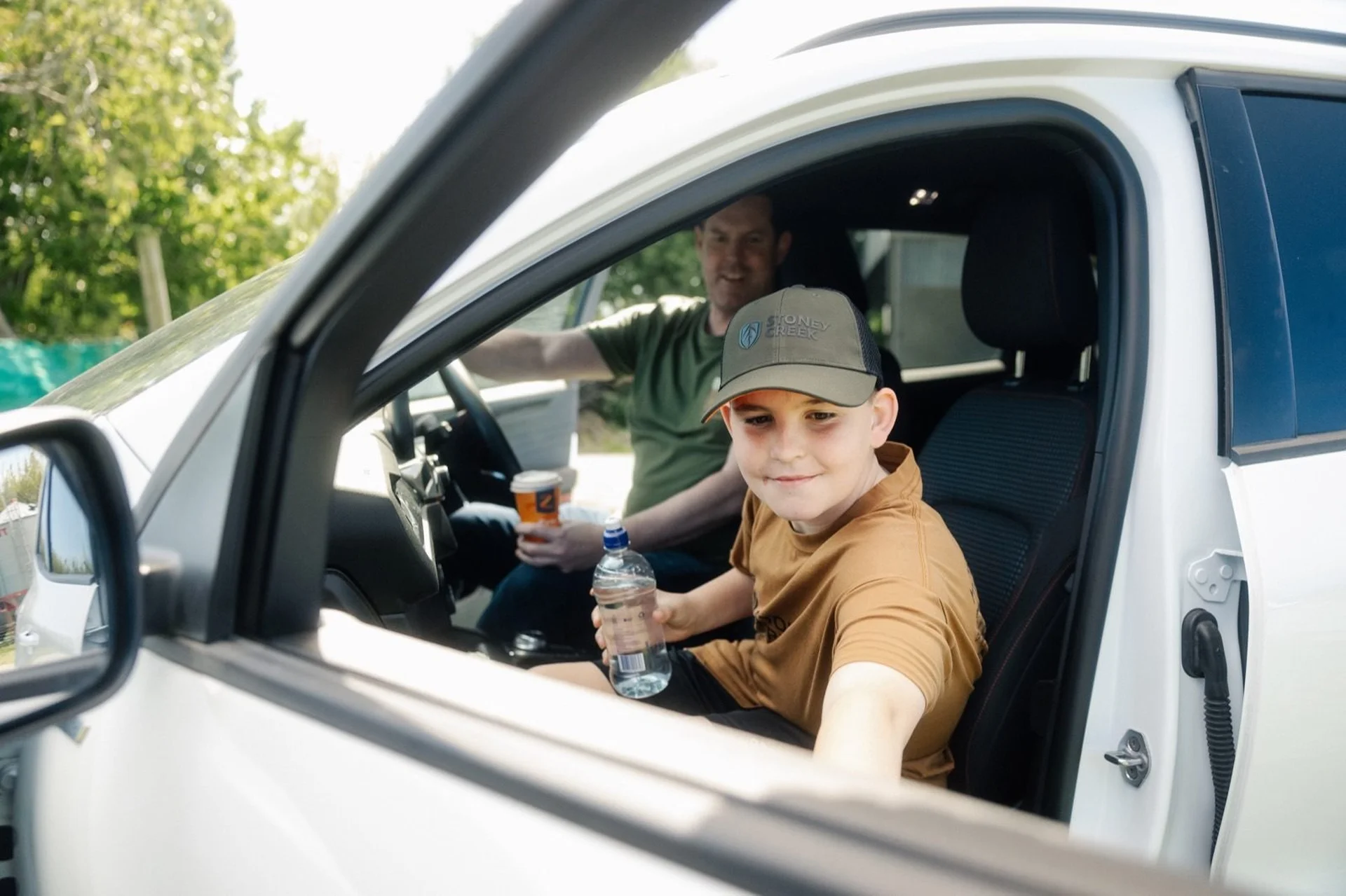 Farmlands Card: A boy in a cap sits in a car's passenger seat holding a water bottle, with a man in the driver's seat.