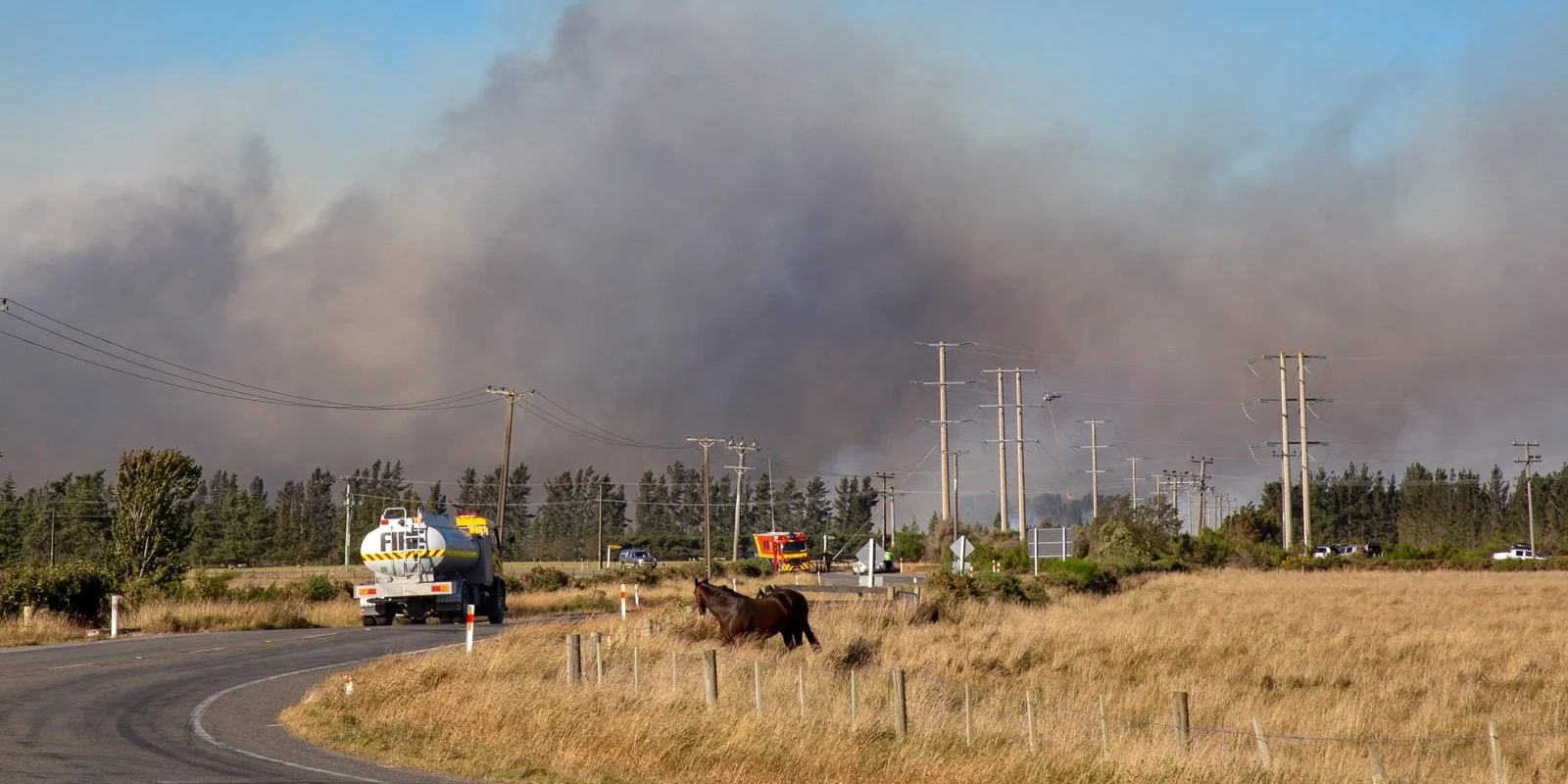Managing wildfire risk on your farm: Smoke billows from a large fire in the distance, with a tanker truck on a curved rural road and fields in the
