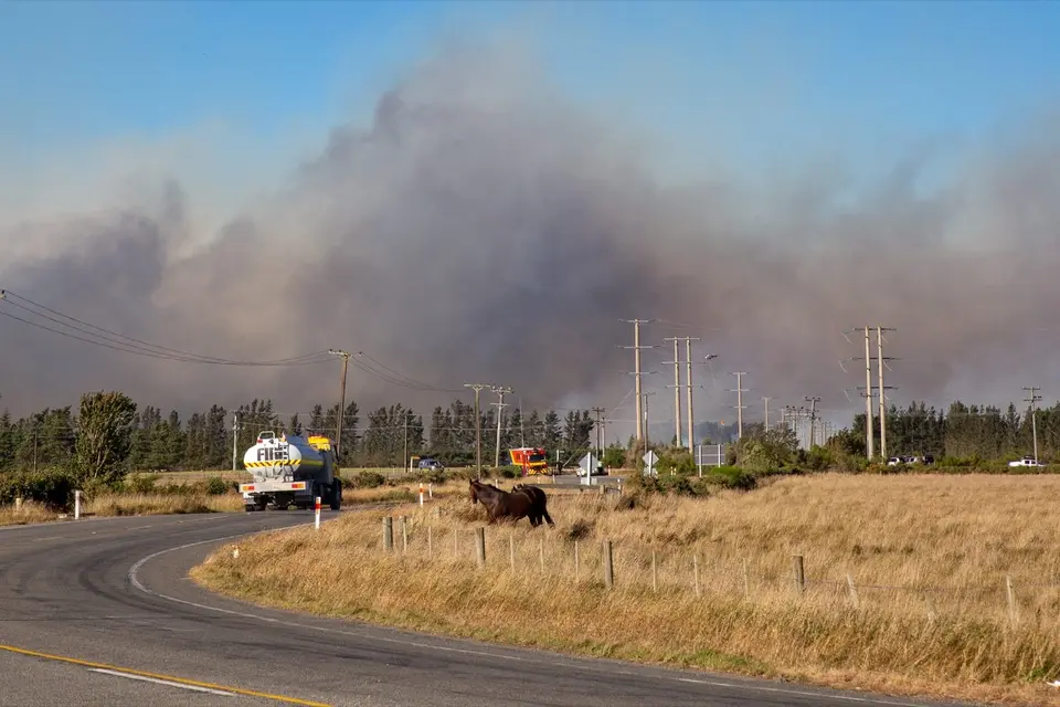 Managing wildfire risk on your farm: Smoke billows from a large fire in the distance, with a tanker truck on a curved rural road and fields in the
