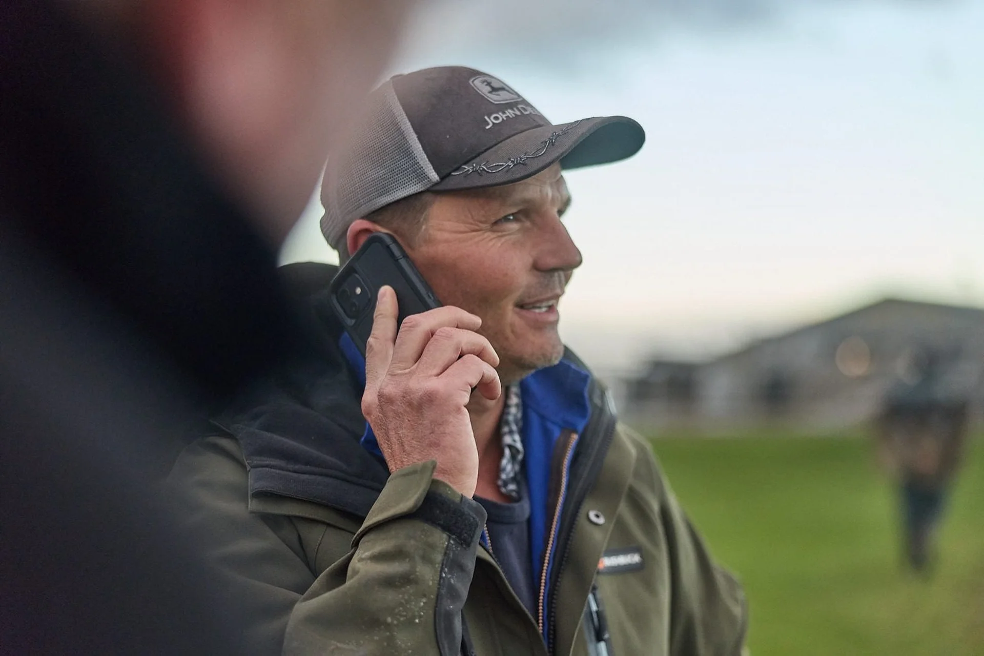 Contact Farmlands: Man in a cap talks on a phone outdoors, smiling, with a grassy field in the blurred background.