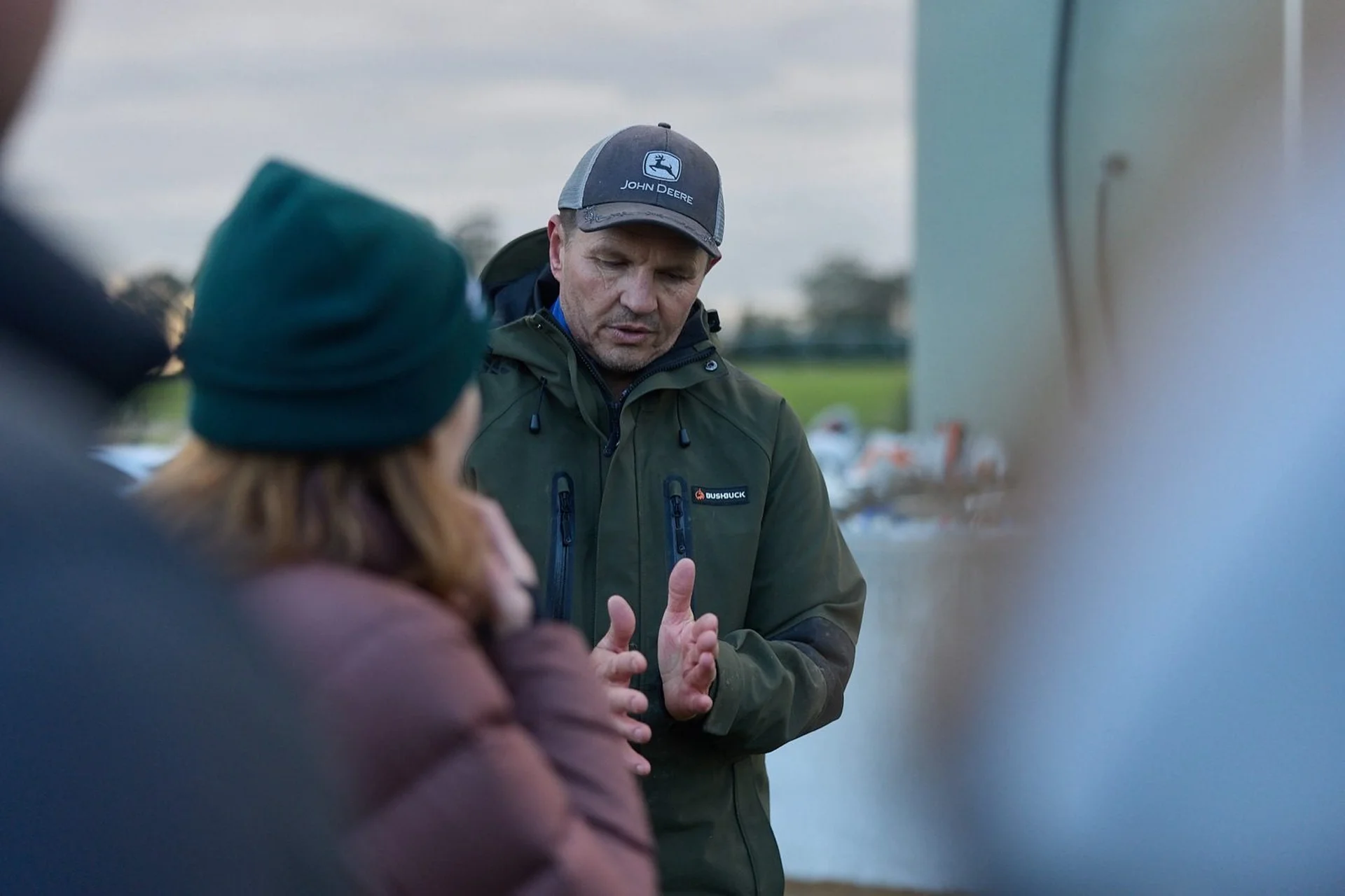 Have your say: A man in a green jacket and cap gestures while talking to a woman outdoors.