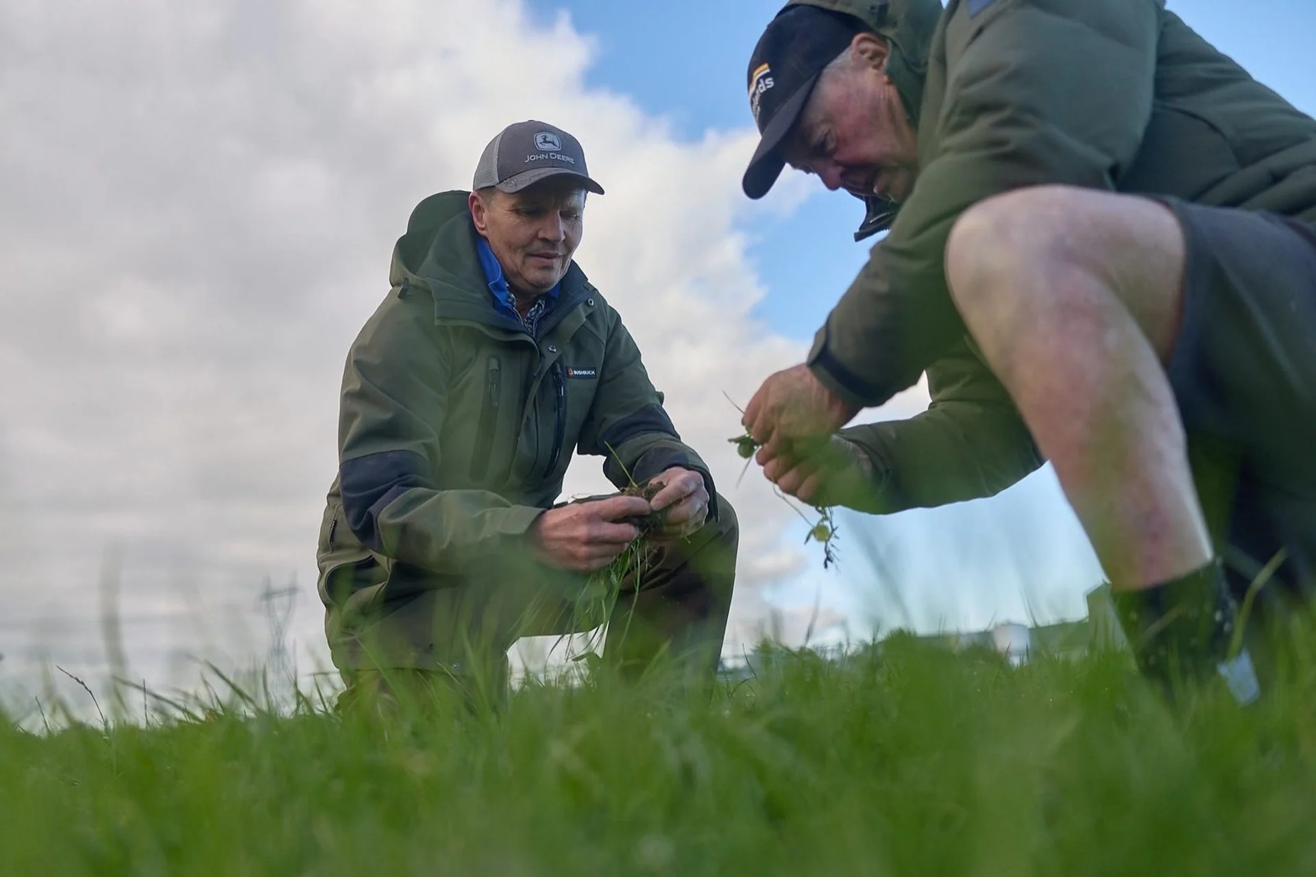 Agronomy: Two people in outdoor clothing examine grass in a field under a partly cloudy sky.