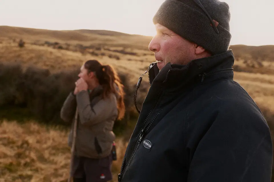 Two people outdoors, one in focus wearing a beanie and jacket, looking ahead; the other blurred in the background, with golden fields behind.
