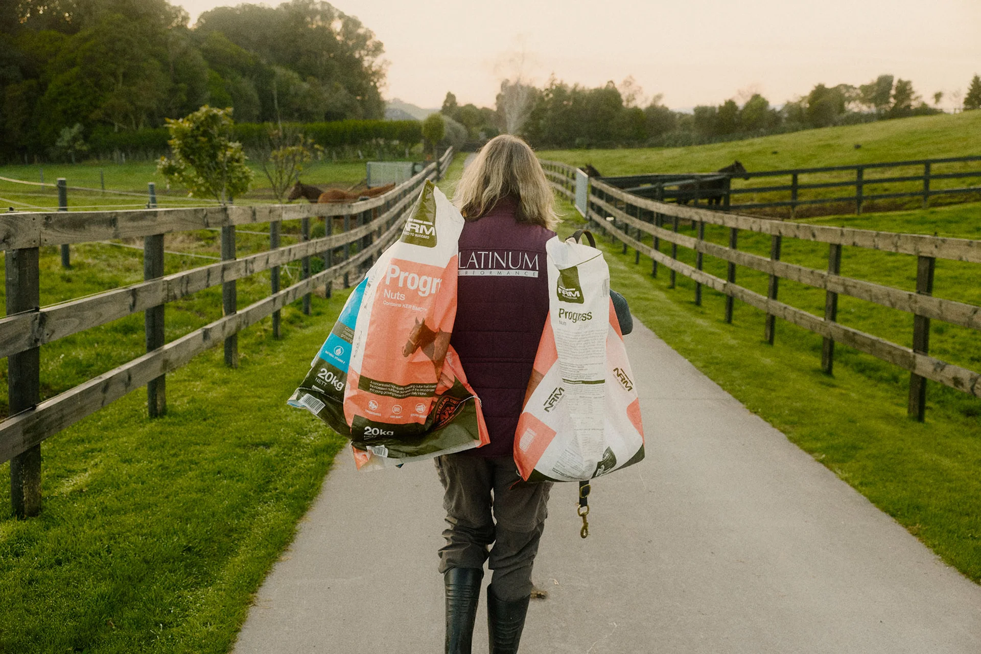 Person walking down a farm path, carrying large NRM feed bags over each shoulder, with horses grazing in the background.