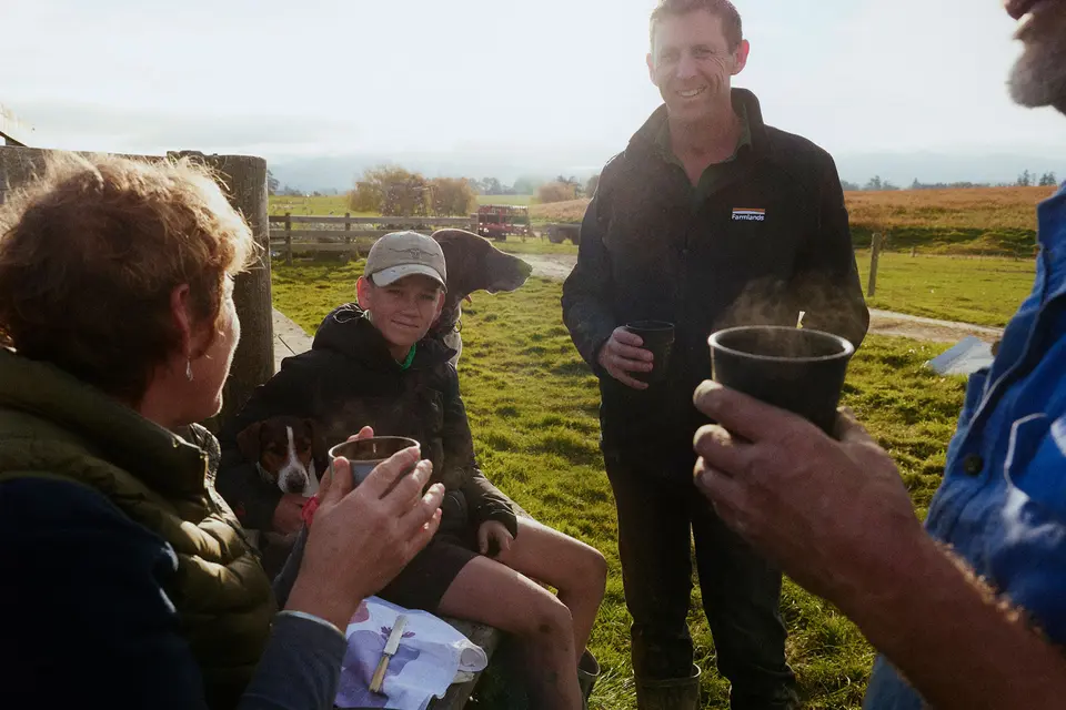 People enjoying hot drinks and conversation outdoors, with a child, a dog, and open countryside in the background.