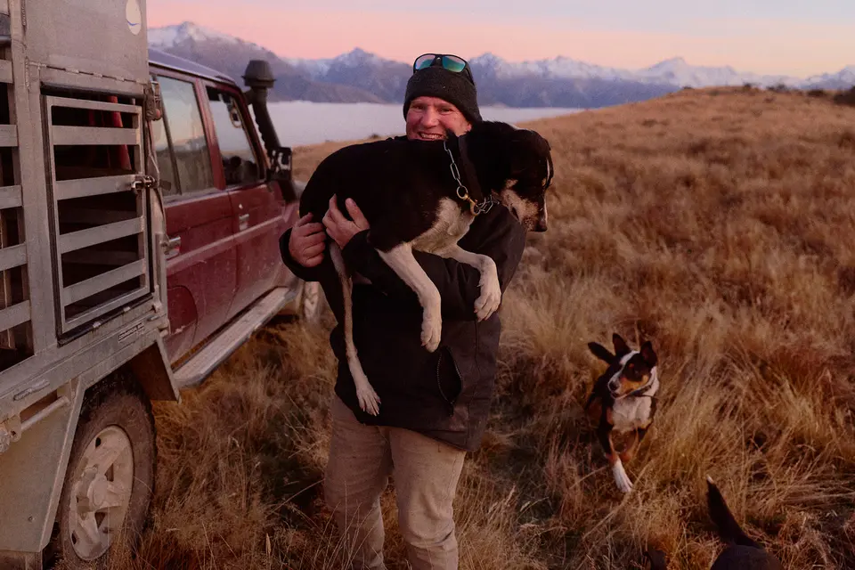 A person holding a dog, standing near a vehicle with cages, in a grassy field with mountains in the background at sunset.
