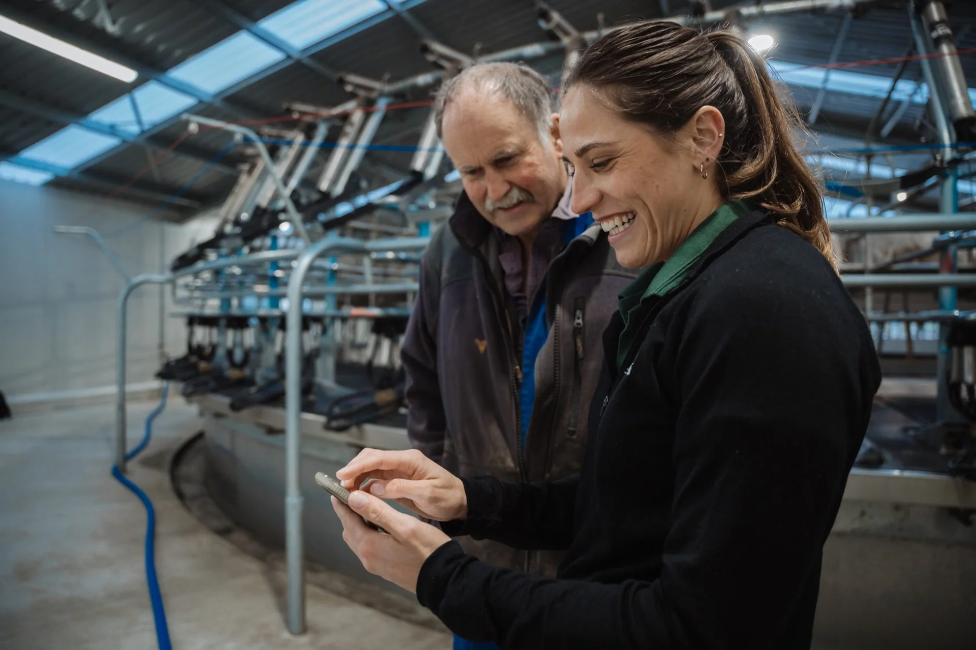 MyFarmlands account: A smiling woman shows her phone to a man in a modern dairy farm setting with milking equipment in the background.