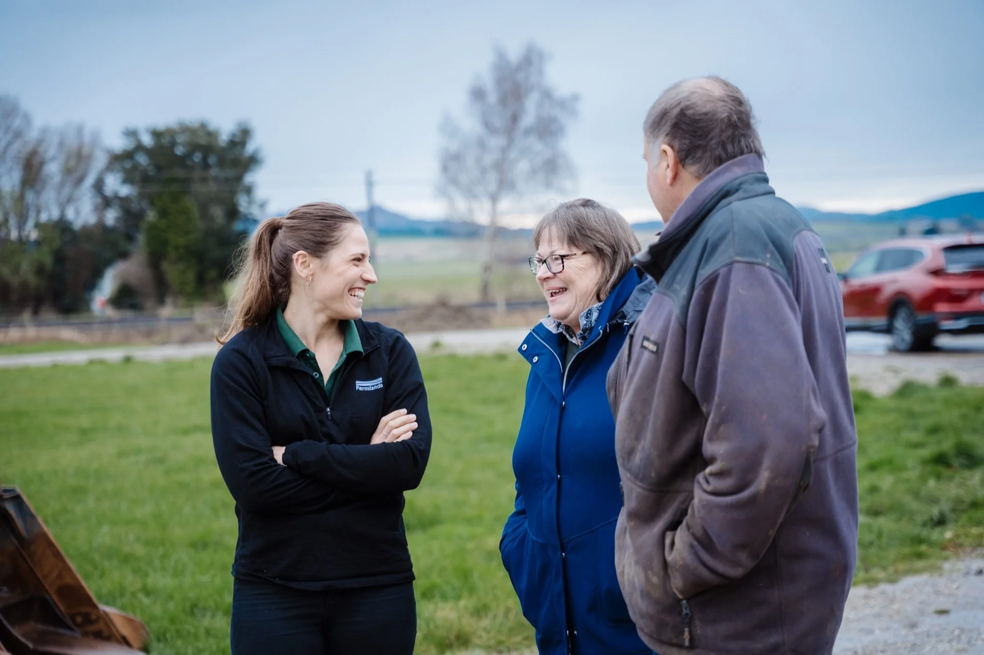 Have your say: Three people talking and smiling outdoors in a rural setting, with fields and a car in the background.