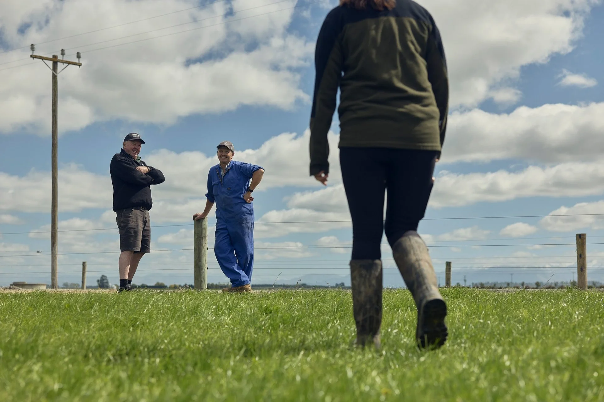 Farmlands agronomy: Three people in casual clothing converse in an open field under a partly cloudy sky. One walks towards two standing by a fence.