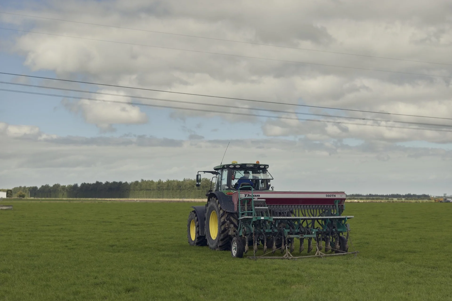 Farmlands fuel: Tractor pulling a seed drill on a vast green field under a cloudy sky, with trees and a distant horizon in the background.