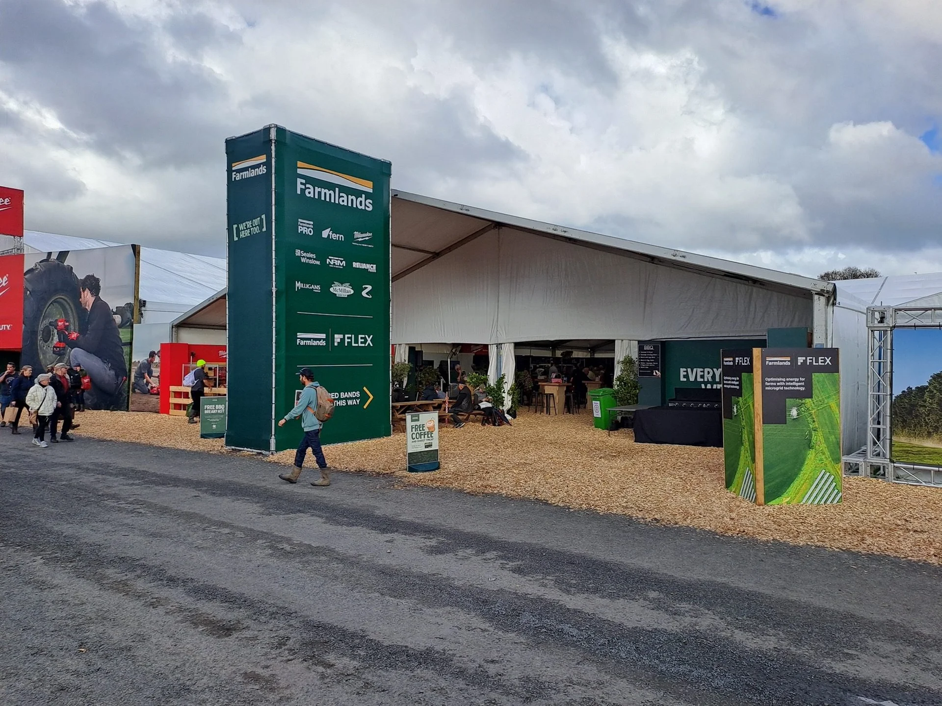 Knowledge and advice: Outdoor event setup with a white marquee tent. Green signage reads "Farmlands" with logos. People walk on gravel path, cloudy