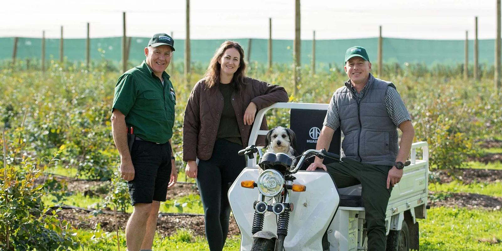 Laurence Frost, Stu Bradbury, Jules Bradbury and their dog stand beside a small white utility vehicle in a field, with a robotic mower in the foreground.