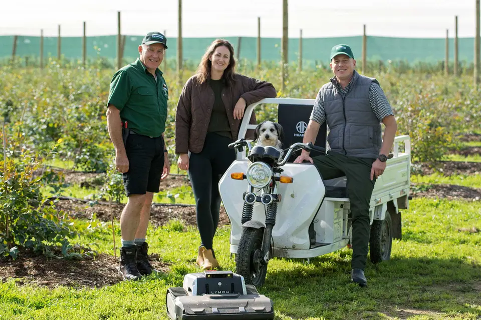 Laurence Frost, Stu Bradbury, Jules Bradbury and their dog stand beside a small white utility vehicle in a field, with a robotic mower in the foreground.