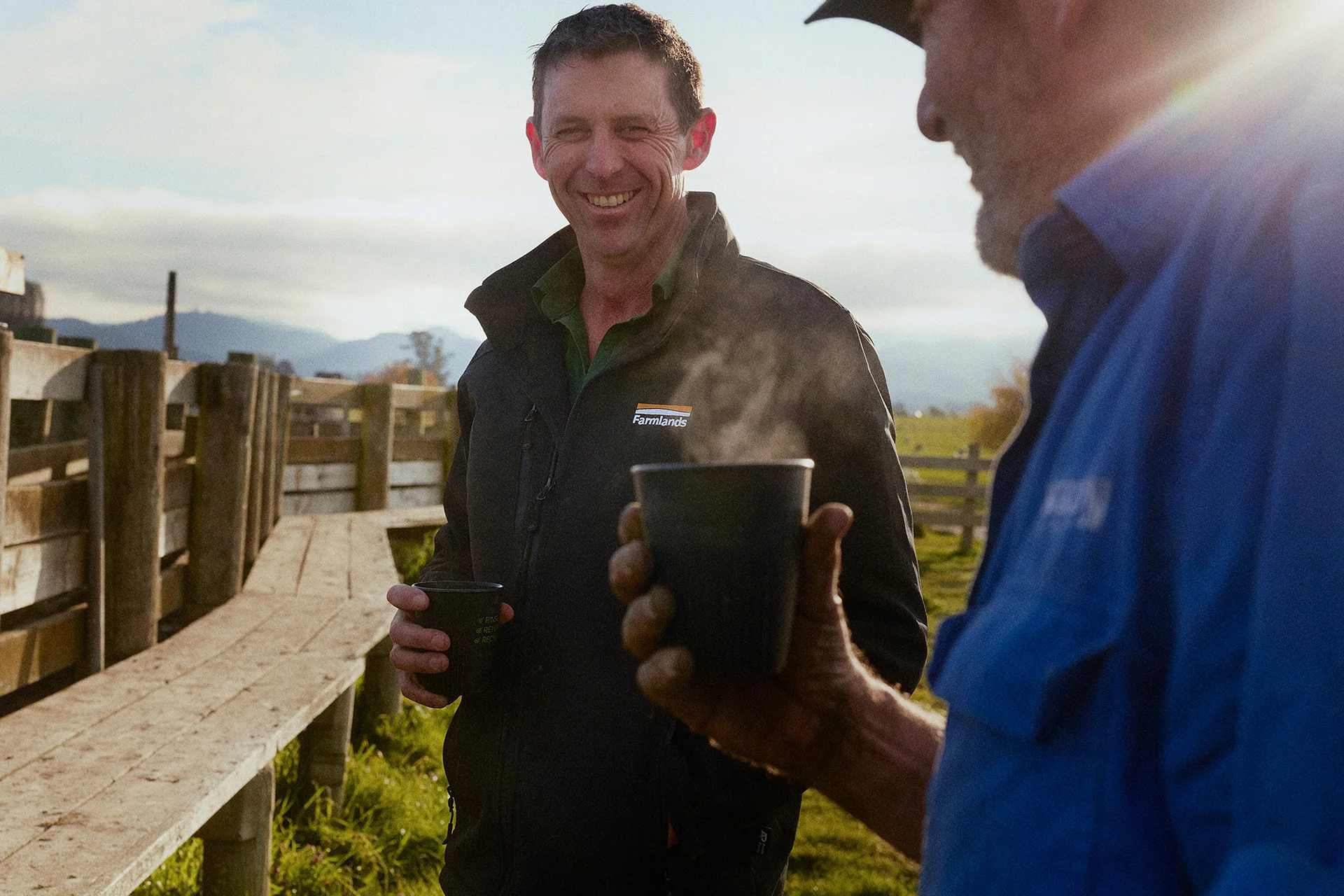 Two men wearing jackets enjoy steaming drinks outdoors on a farm, one is wearing a Farmlands jacket