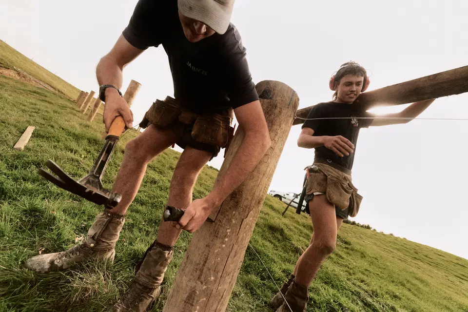 Two workers build a fence, one uses a hammer while the other holds the wire. They are in a grassy field, wearing tool belts and boots.