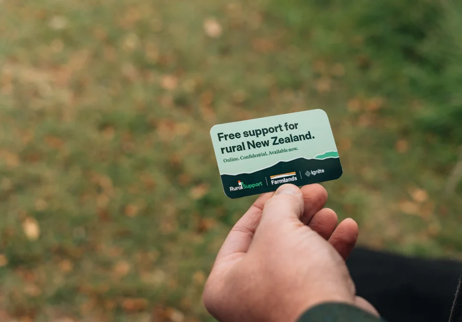 Hand holding a card with "Free support for rural New Zealand" printed on it, against a grassy outdoor background.