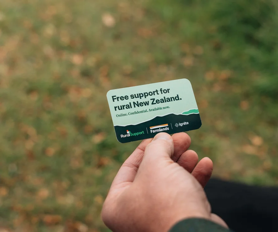 Hand holding a card with "Free support for rural New Zealand" printed on it, against a grassy outdoor background.