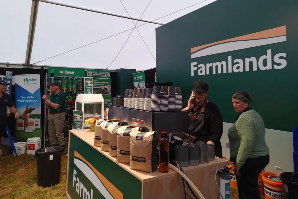 Inside a tent, a coffee stall with Farmlands branding, coffee cups, and patrons. Nearby, promotional banners and equipment are displayed.