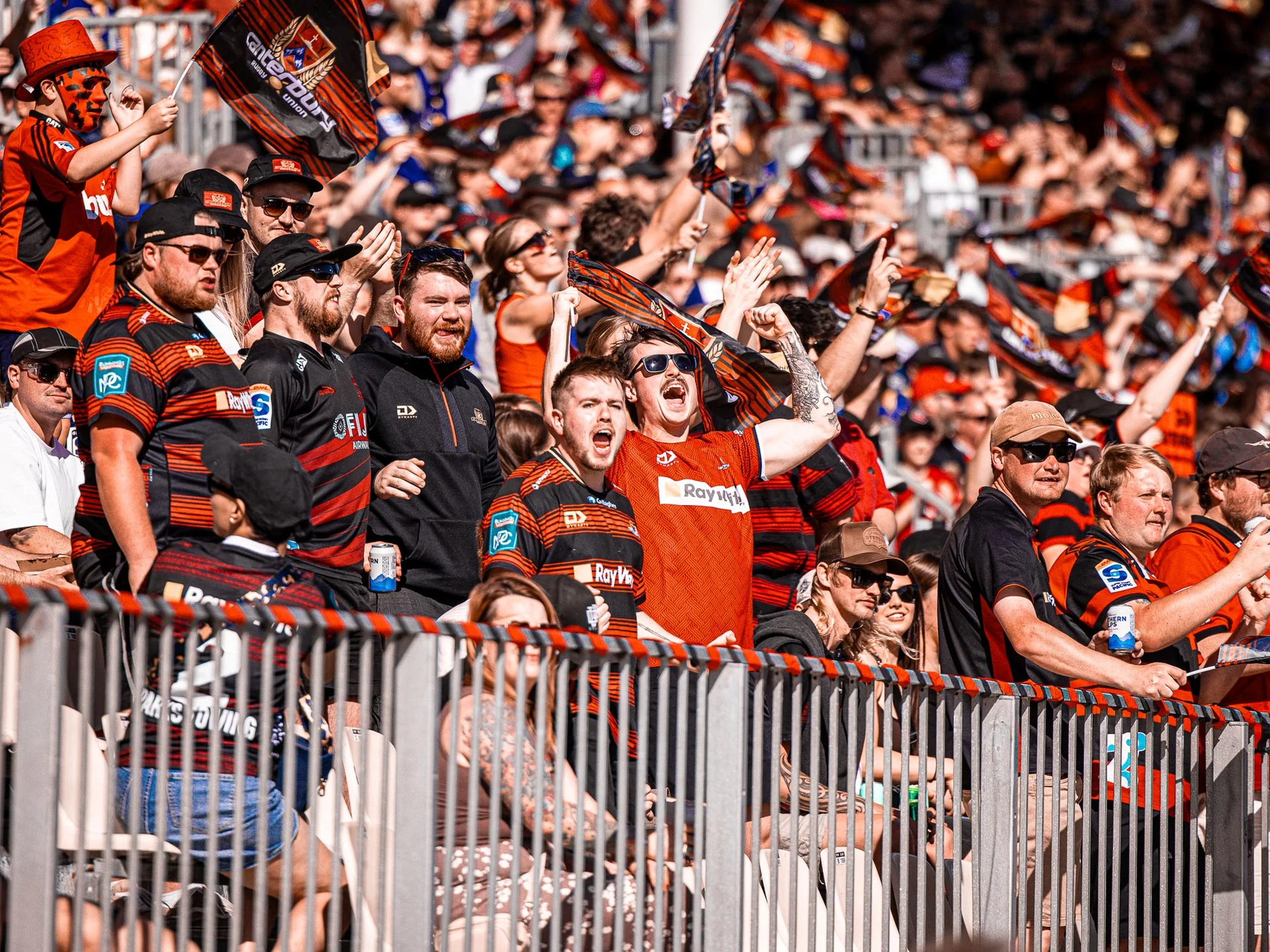 Cheering sports fans wearing red and black jerseys, waving flags and raising their hands, packed in stadium stands.