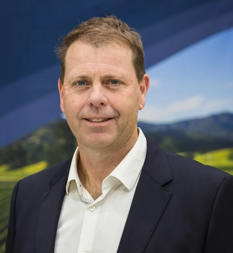 Farmlands team: Man in a suit and white shirt smiling, standing in front of a scenic backdrop featuring hills and fields under a clear blue sky.